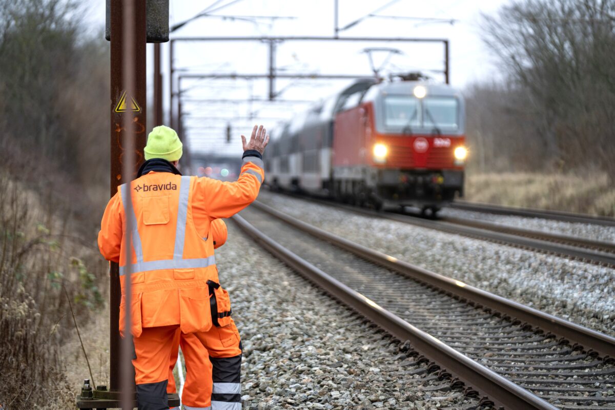 En mand i arbejdstøj fra Bravida vinker til et tog der kører forbi ham.